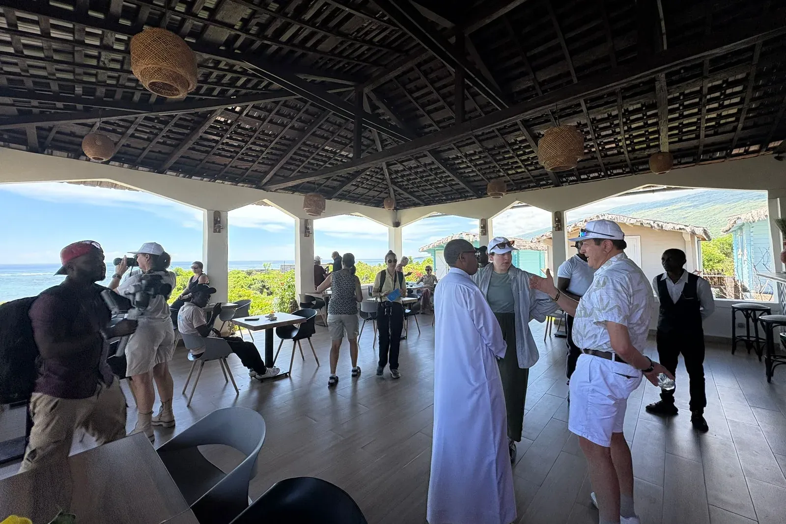 Un groupe de voyageurs et leurs accompagnateurs discutent sous la charpente de bois d'une terrasse ouverte sur la mer, aux Comores.