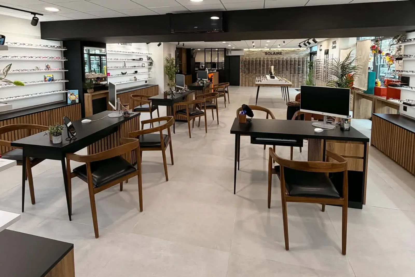Vue d'ensemble du magasin : des tables noires avec leurs chaises en bois, des murs de montures sur des étagères blanches, et un espace solaire au fond.