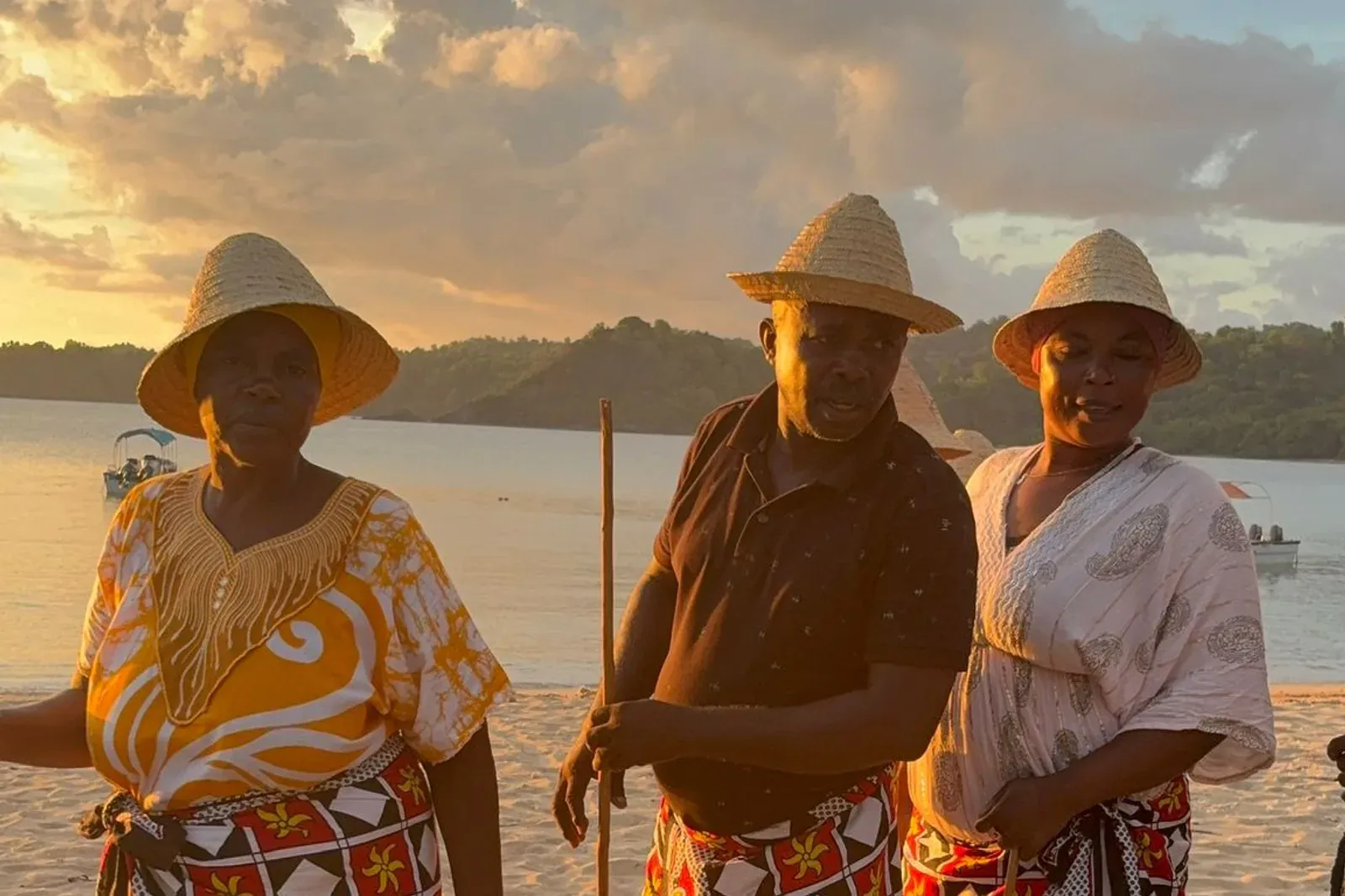 Trois habitants des Comores en tenue traditionnelle, chapeau de paille et pagne à motifs, sur une plage de sable clair au coucher du soleil.