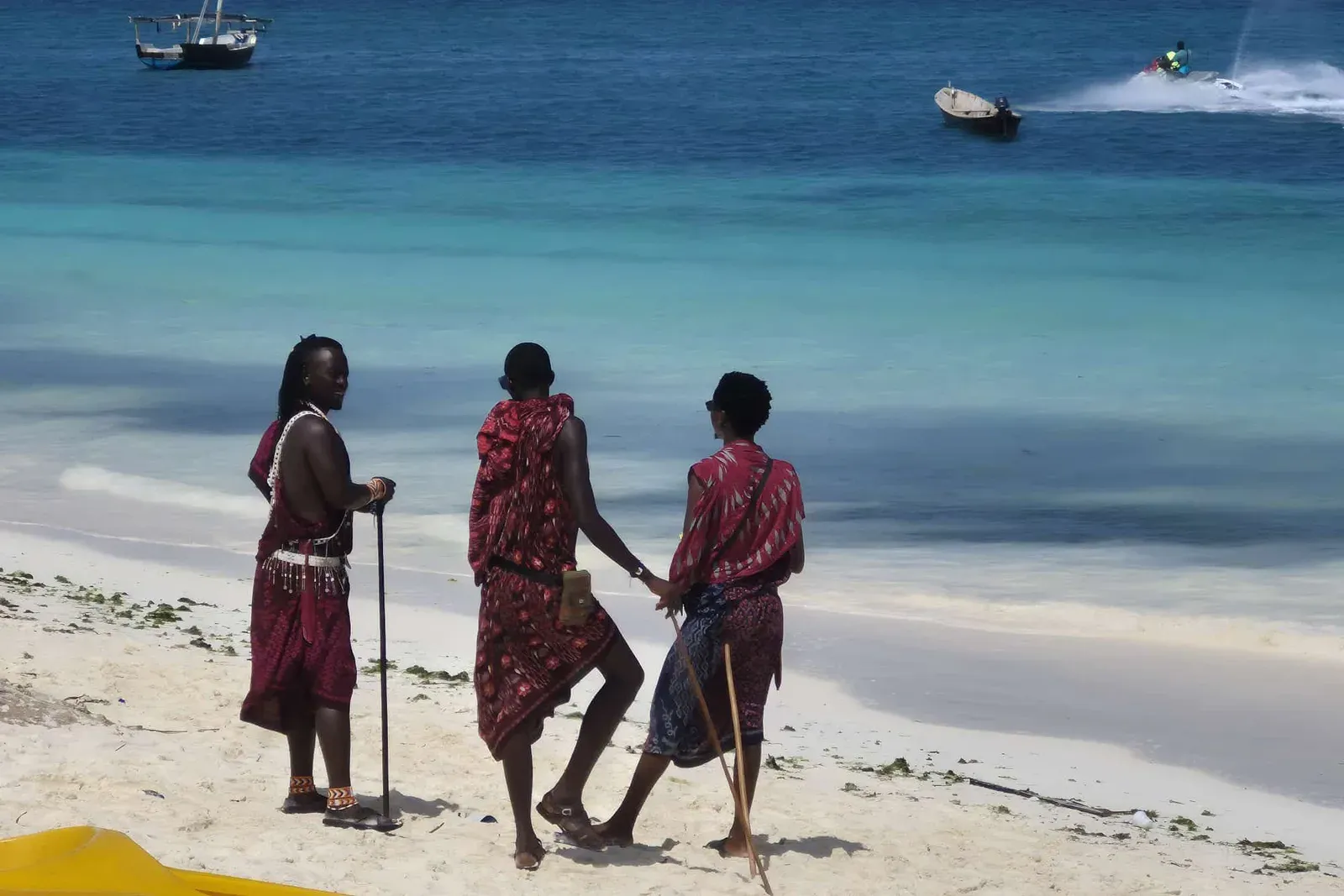 Trois hommes en tenue rouge marchent sur le sable blanc d'une plage de Zanzibar, devant une mer turquoise où mouillent des barques.
